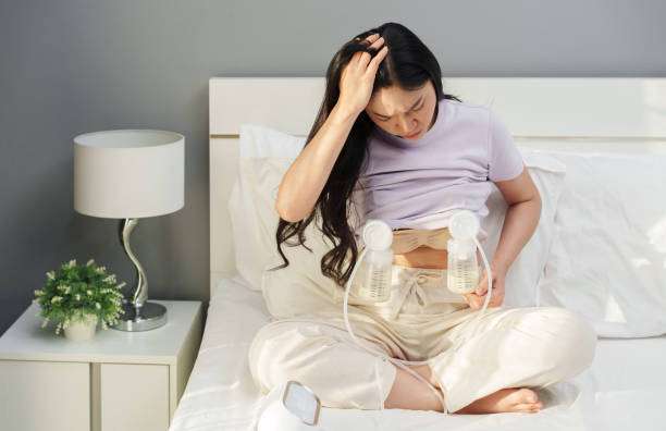 Asian mother using breast pump, looking tired on bed.