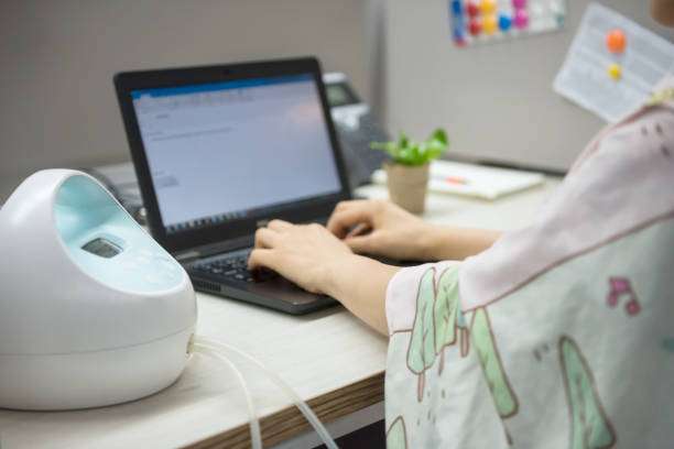 Woman working on laptop at desk while using an electric breast pump.